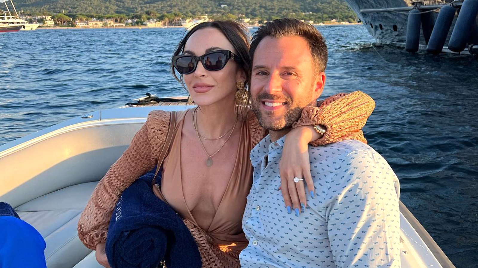 Farrah Aldjufrie in deep V neck beige dress and black sunglasses puts her arm around Alex Manos wearing a button down while riding on a boat with water in background